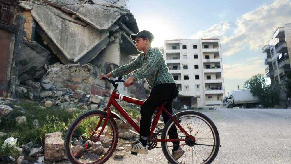 A boy riding a bicycle past rubble and damaged buildings